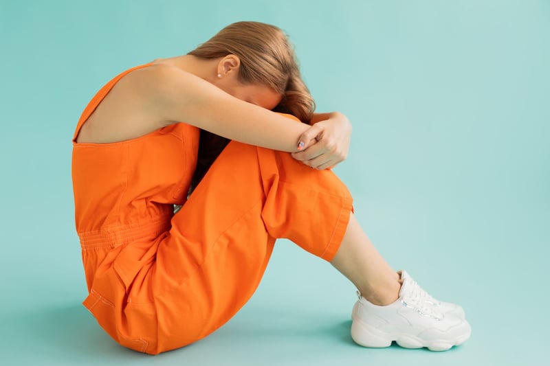 wanita pirang dengan baju oranye dan sepatu kets putih memeluk lutut dan menyembunyikan wajah